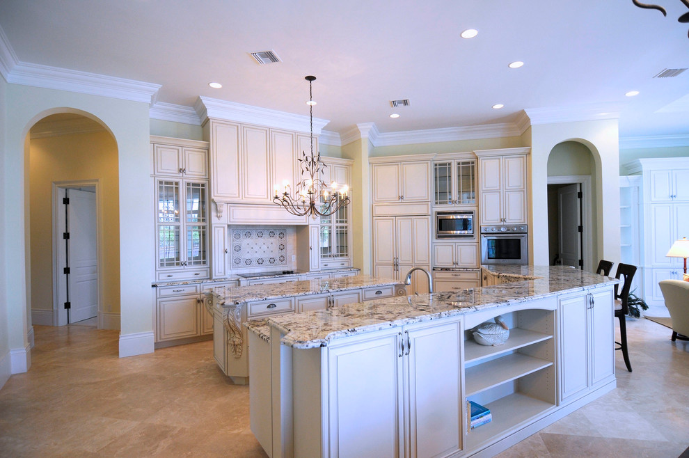 Inspiration for a traditional l-shaped kitchen/diner in Miami with a belfast sink, beige cabinets, granite worktops, stainless steel appliances, marble flooring, raised-panel cabinets, multiple islands and beige floors.