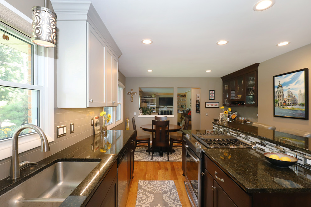This is an example of a medium sized classic l-shaped kitchen/diner in Grand Rapids with a double-bowl sink, recessed-panel cabinets, dark wood cabinets, granite worktops, beige splashback, metro tiled splashback, stainless steel appliances, light hardwood flooring, an island and brown floors.