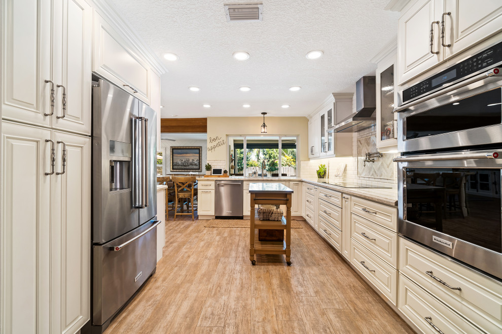 Klassische Küche in U-Form mit profilierten Schrankfronten, beigen Schränken, Küchenrückwand in Weiß, Küchengeräten aus Edelstahl, braunem Holzboden, Kücheninsel, braunem Boden und grauer Arbeitsplatte in Miami