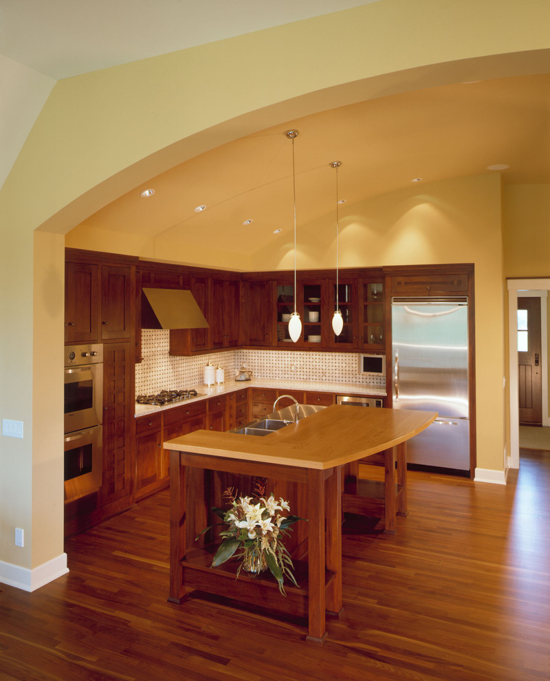 ミネアポリスにあるトラディショナルスタイルのおしゃれなL型キッチン (ダブルシンク、シェーカースタイル扉のキャビネット、中間色木目調キャビネット、白いキッチンパネル、シルバーの調理設備) の写真
