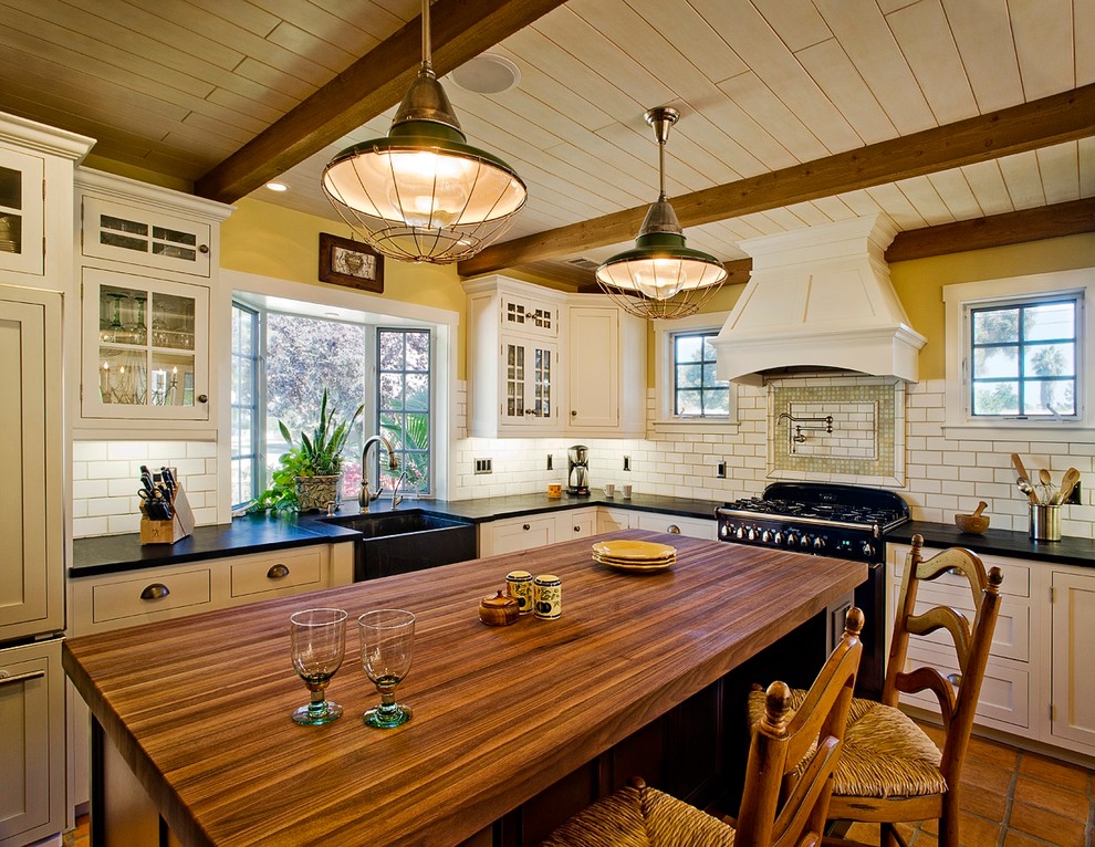 Example of a large classic l-shaped terra-cotta tile enclosed kitchen design in Santa Barbara with a farmhouse sink, shaker cabinets, white cabinets, wood countertops, white backsplash, subway tile backsplash, black appliances and an island