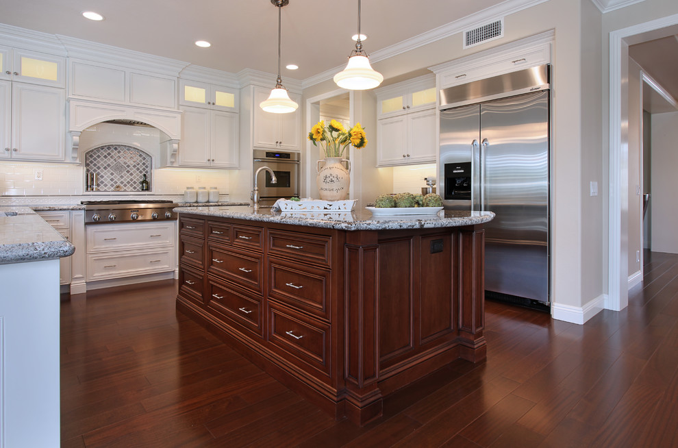 Inspiration for a large timeless u-shaped medium tone wood floor eat-in kitchen remodel in Orange County with a single-bowl sink, raised-panel cabinets, white cabinets, granite countertops, white backsplash, ceramic backsplash, stainless steel appliances and an island
