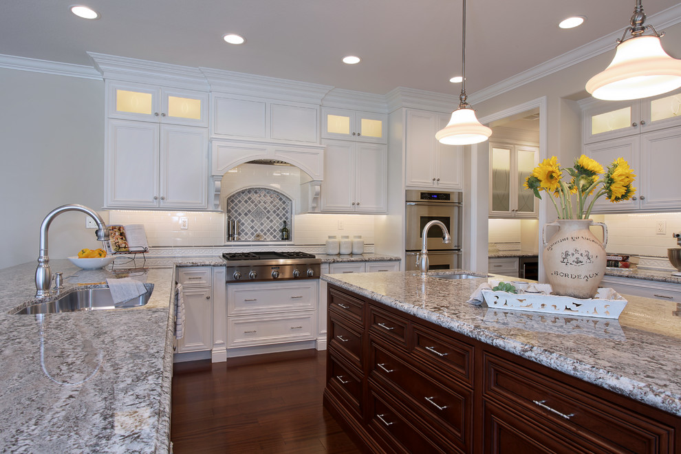 Example of a large classic u-shaped medium tone wood floor eat-in kitchen design in Orange County with a single-bowl sink, raised-panel cabinets, white cabinets, granite countertops, white backsplash, ceramic backsplash, stainless steel appliances and an island