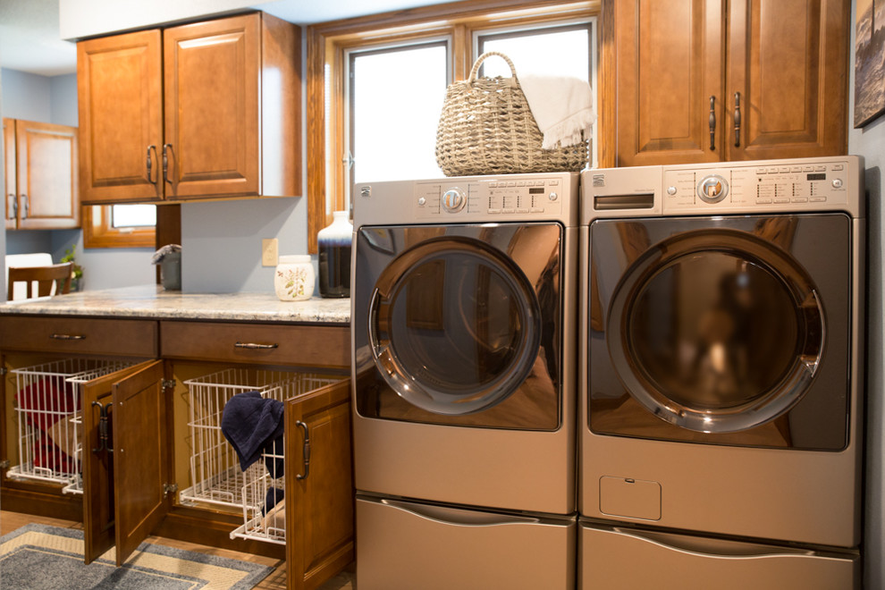 Inspiration for a traditional single-wall separated utility room in Cedar Rapids with granite worktops, medium hardwood flooring, raised-panel cabinets, medium wood cabinets, blue walls, a stacked washer and dryer and brown floors.