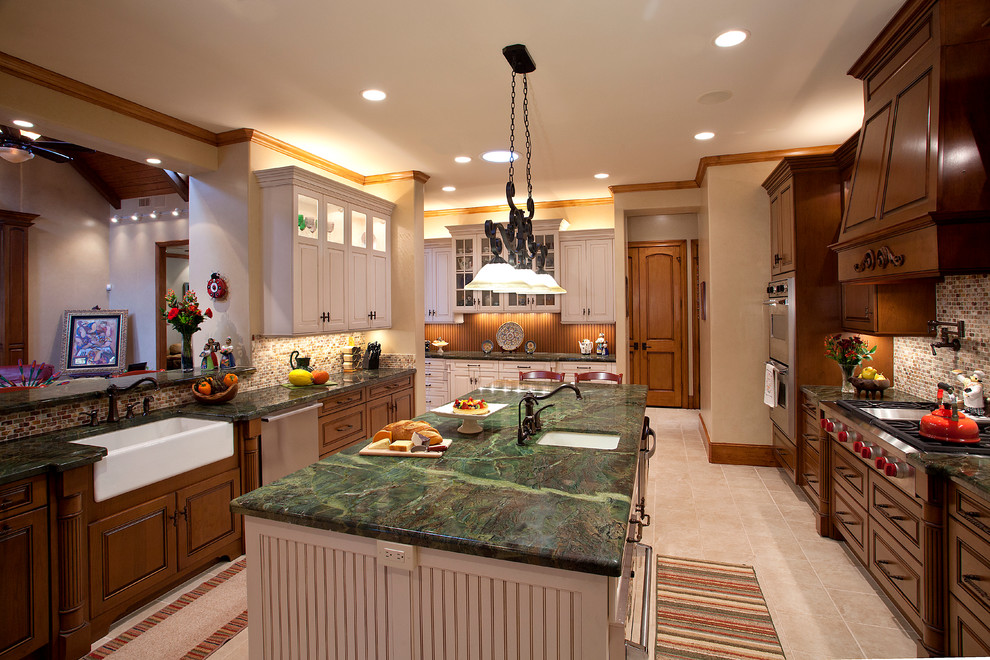 Large traditional galley kitchen/diner in Houston with a belfast sink, raised-panel cabinets, granite worktops, stainless steel appliances, brown cabinets, beige splashback, an island, beige floors and green worktops.