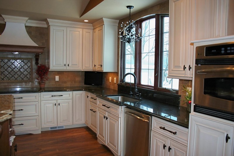 Example of a mid-sized classic l-shaped medium tone wood floor and brown floor eat-in kitchen design in Cedar Rapids with an undermount sink, raised-panel cabinets, white cabinets, granite countertops, beige backsplash, stone tile backsplash, stainless steel appliances and an island