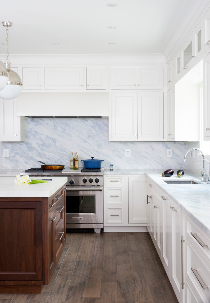 Klassisk inredning av ett l-kök, med en undermonterad diskho, luckor med infälld panel, vita skåp, rostfria vitvaror, en köksö, flerfärgad stänkskydd och mörkt trägolv