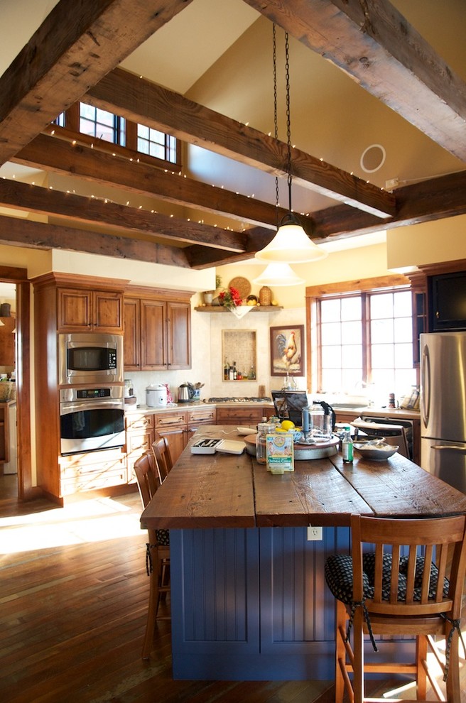 This is an example of a large rustic l-shaped enclosed kitchen in Other with raised-panel cabinets, medium wood cabinets, an island, composite countertops, stainless steel appliances, a built-in sink and medium hardwood flooring.