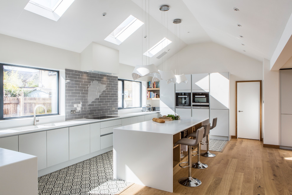 Moderne Küche in U-Form mit Unterbauwaschbecken, flächenbündigen Schrankfronten, weißen Schränken, Küchenrückwand in Grau, Elektrogeräten mit Frontblende, braunem Holzboden, Kücheninsel, braunem Boden und weißer Arbeitsplatte in Oxfordshire