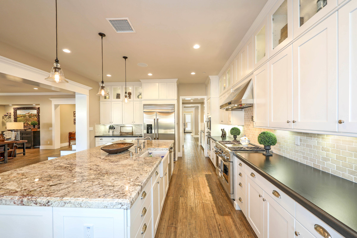 Inspiration for a large traditional l-shaped open plan kitchen in Orange County with a belfast sink, shaker cabinets, white cabinets, engineered stone countertops, beige splashback, metro tiled splashback, stainless steel appliances, medium hardwood flooring, an island and brown floors.