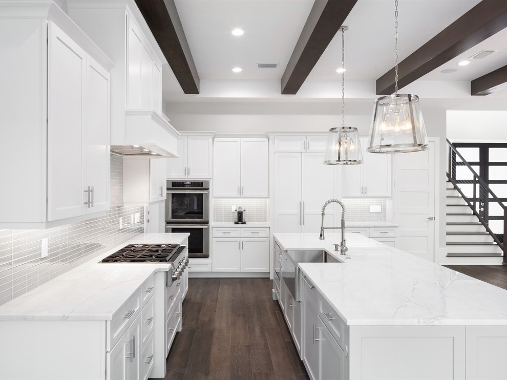 Large traditional l-shaped open plan kitchen in Orlando with a belfast sink, shaker cabinets, white cabinets, grey splashback, glass tiled splashback, stainless steel appliances, dark hardwood flooring, an island, brown floors, white worktops and engineered stone countertops.