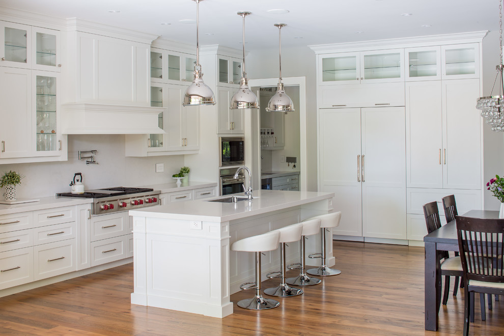 Large traditional l-shaped kitchen/diner in Toronto with a submerged sink, shaker cabinets, white cabinets, engineered stone countertops, white splashback, stone slab splashback, integrated appliances, medium hardwood flooring and an island.
