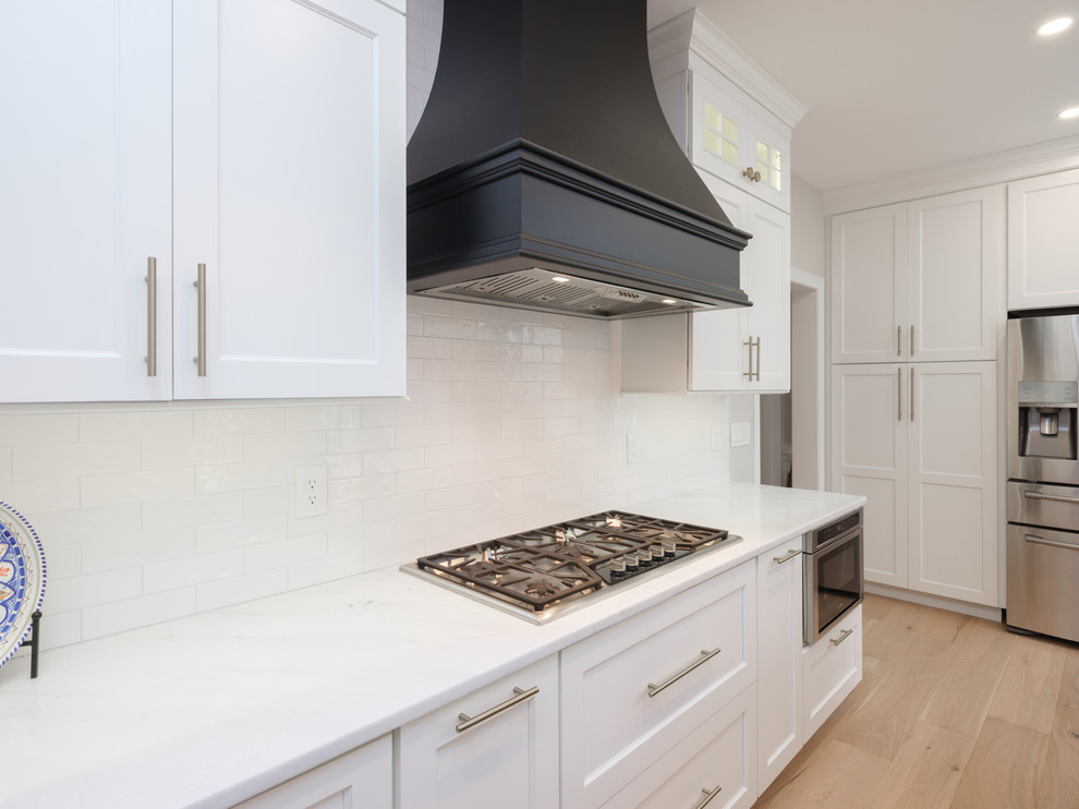 Example of a large transitional light wood floor kitchen design in Philadelphia with an undermount sink, shaker cabinets, white cabinets, quartz countertops, white backsplash, porcelain backsplash, stainless steel appliances, an island and white countertops