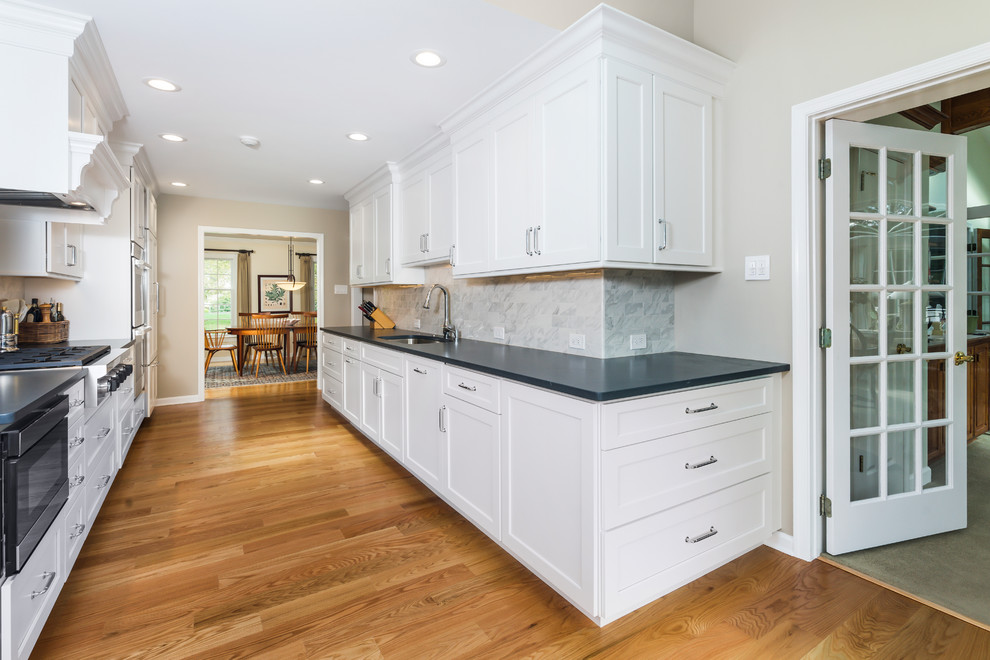 Mid-sized transitional galley medium tone wood floor and brown floor enclosed kitchen photo in Philadelphia with an undermount sink, flat-panel cabinets, white cabinets, quartz countertops, gray backsplash, porcelain backsplash, paneled appliances, no island and black countertops