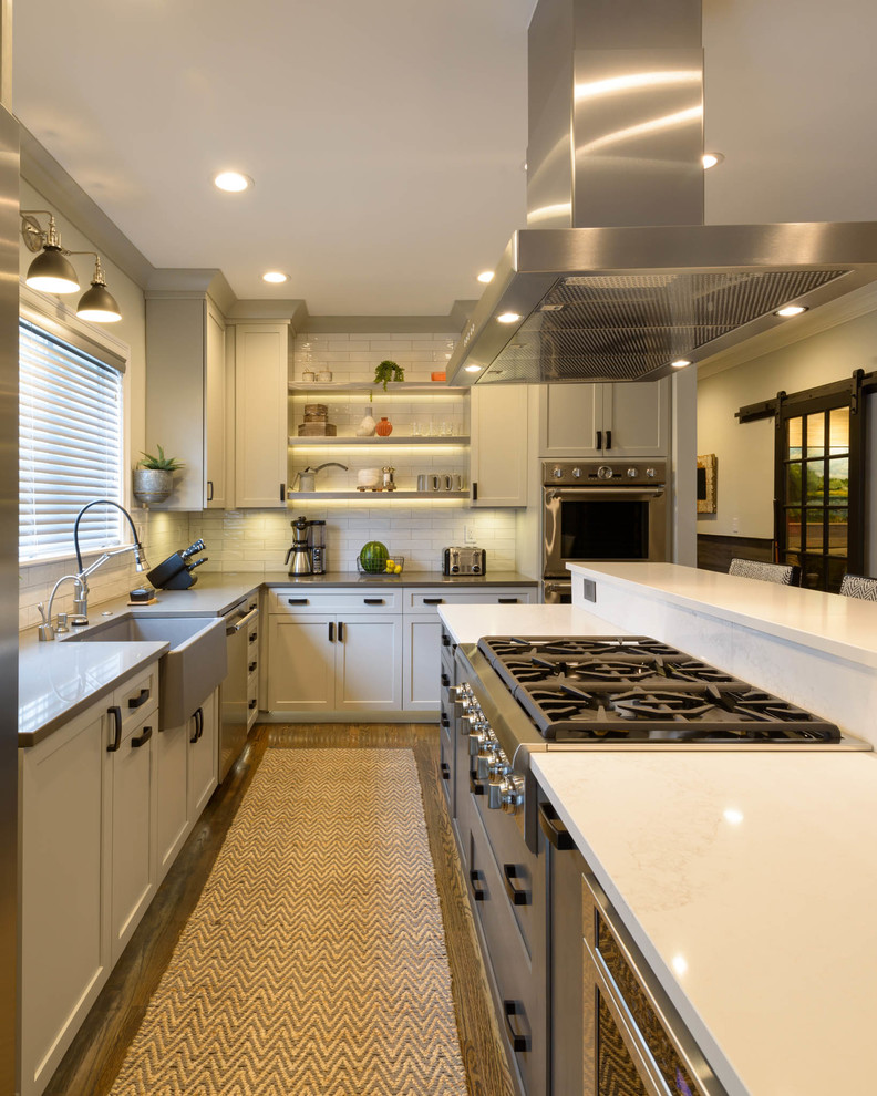 This is an example of a medium sized classic l-shaped kitchen/diner in Boston with a belfast sink, shaker cabinets, white cabinets, white splashback, metro tiled splashback, stainless steel appliances, medium hardwood flooring, an island and brown floors.