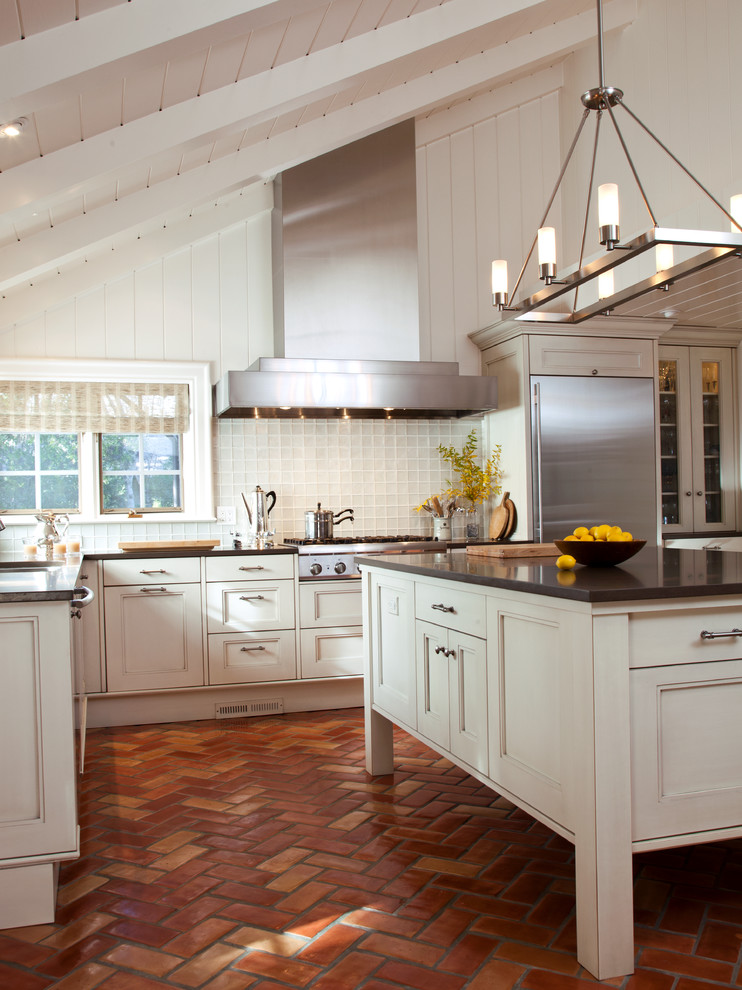Bild på ett mellanstort vintage kök, med luckor med infälld panel, vita skåp, bänkskiva i kvarts, vitt stänkskydd, stänkskydd i glaskakel, rostfria vitvaror, tegelgolv och en köksö