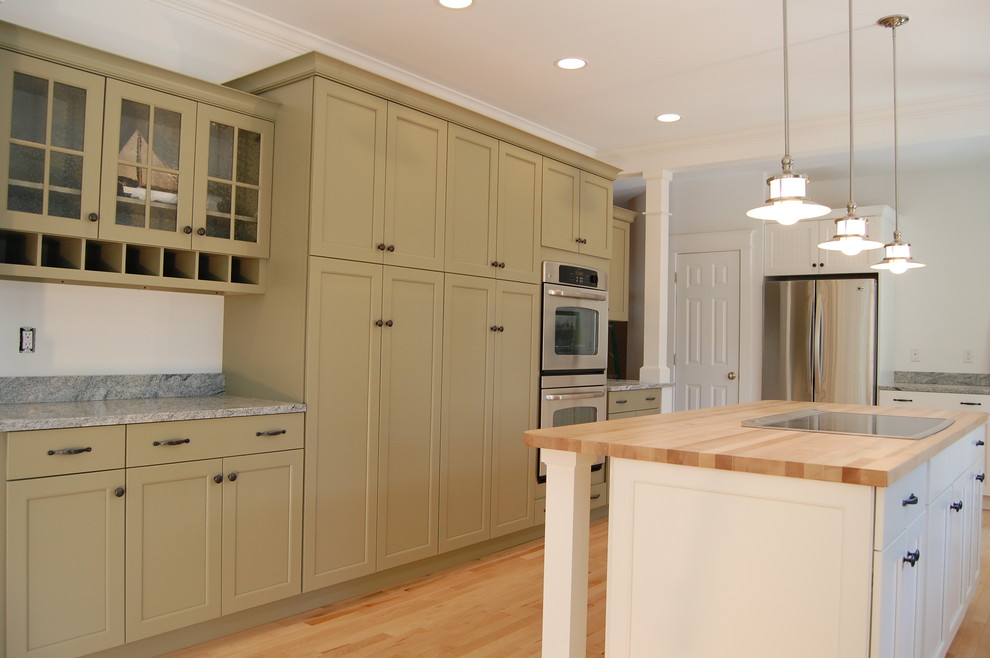 This is an example of an expansive rural u-shaped kitchen/diner in Portland Maine with a belfast sink, shaker cabinets, green cabinets, granite worktops, grey splashback, stone slab splashback, stainless steel appliances, light hardwood flooring and an island.