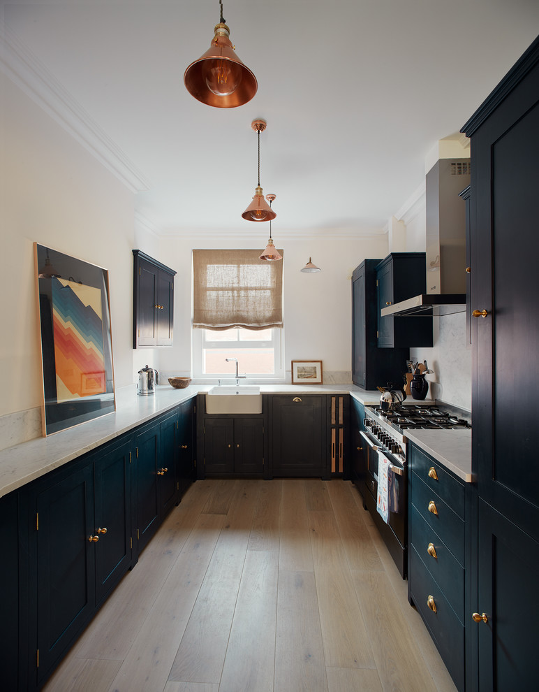 This is an example of a large traditional u-shaped open plan kitchen in London with a belfast sink, shaker cabinets, blue cabinets, marble worktops, white splashback, marble splashback, black appliances, light hardwood flooring, no island, grey floors and white worktops.