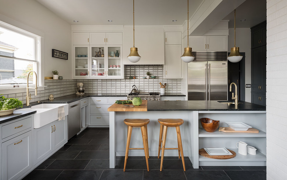 Design ideas for a traditional l-shaped kitchen in Seattle with a belfast sink, glass-front cabinets, beige cabinets, beige splashback, stainless steel appliances, an island, black floors and black worktops.