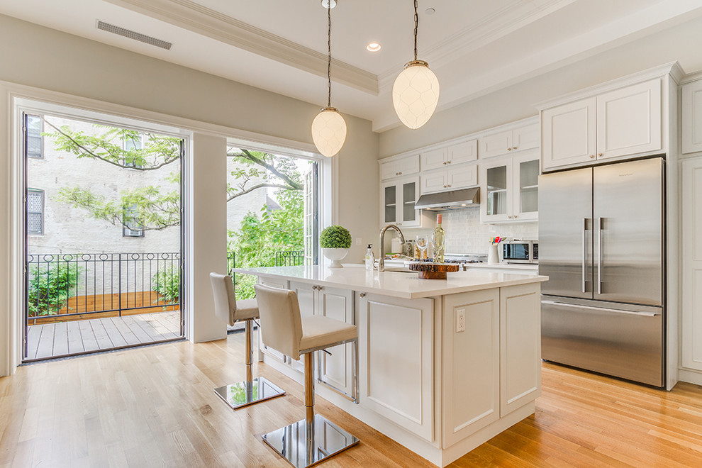 Example of a transitional light wood floor kitchen design in New York with an undermount sink, recessed-panel cabinets, white cabinets, white backsplash, subway tile backsplash, stainless steel appliances, an island and white countertops