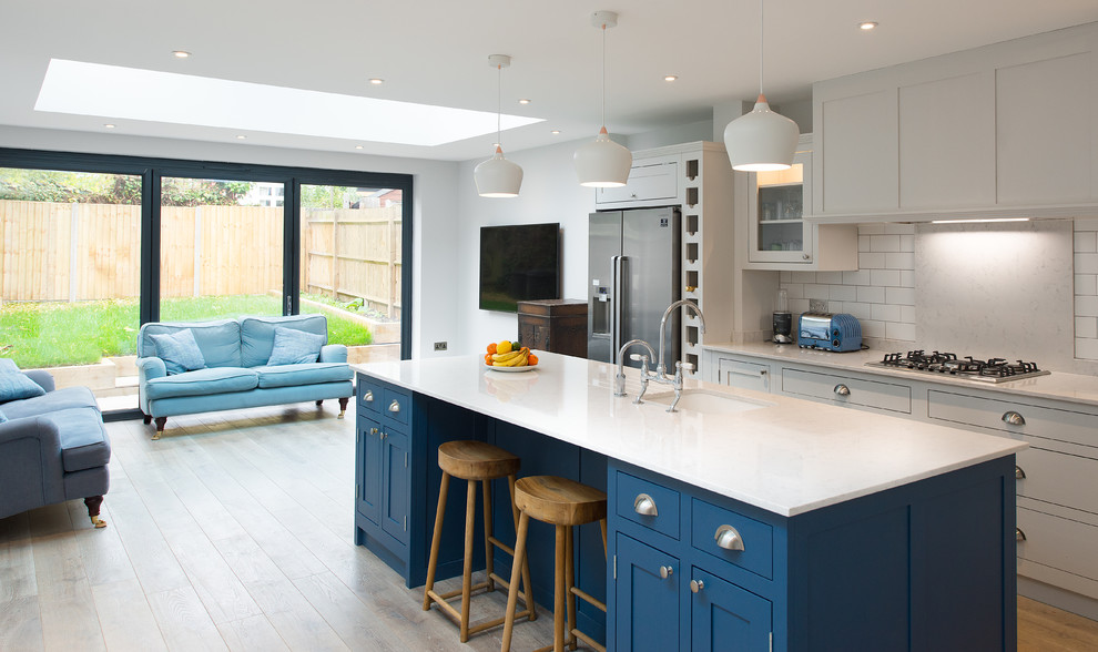 This is an example of a medium sized traditional open plan kitchen in London with shaker cabinets, marble worktops, white splashback, metro tiled splashback, stainless steel appliances, light hardwood flooring, an island, a submerged sink and blue cabinets.