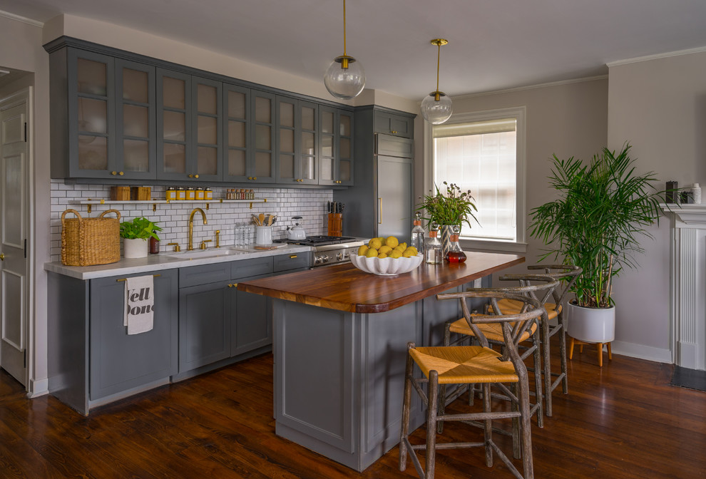 Inspiration for a transitional single-wall dark wood floor and brown floor kitchen remodel in Boston with an undermount sink, gray cabinets, wood countertops, white backsplash, subway tile backsplash, paneled appliances, an island and glass-front cabinets