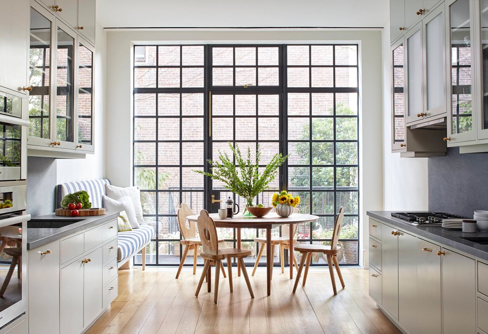 Inspiration for a mid-sized transitional galley medium tone wood floor eat-in kitchen remodel in New York with glass-front cabinets, white cabinets, gray backsplash, no island, solid surface countertops, stone slab backsplash, stainless steel appliances, an undermount sink and gray countertops