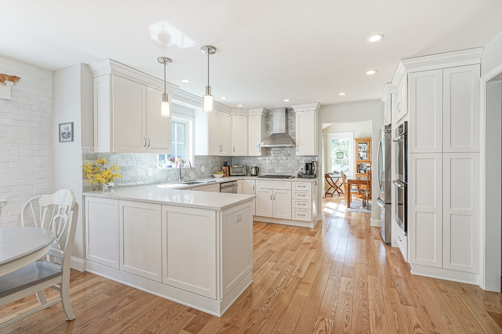 Modelo de cocina clásica renovada de tamaño medio con puertas de armario blancas, electrodomésticos de acero inoxidable, suelo de madera clara, suelo marrón y encimeras blancas