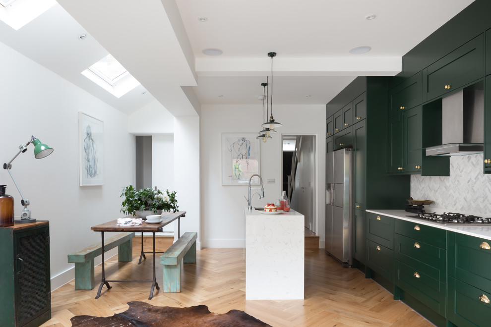 Design ideas for a traditional galley kitchen/diner in London with a submerged sink, shaker cabinets, green cabinets, white splashback, stainless steel appliances, medium hardwood flooring, an island and brown floors.