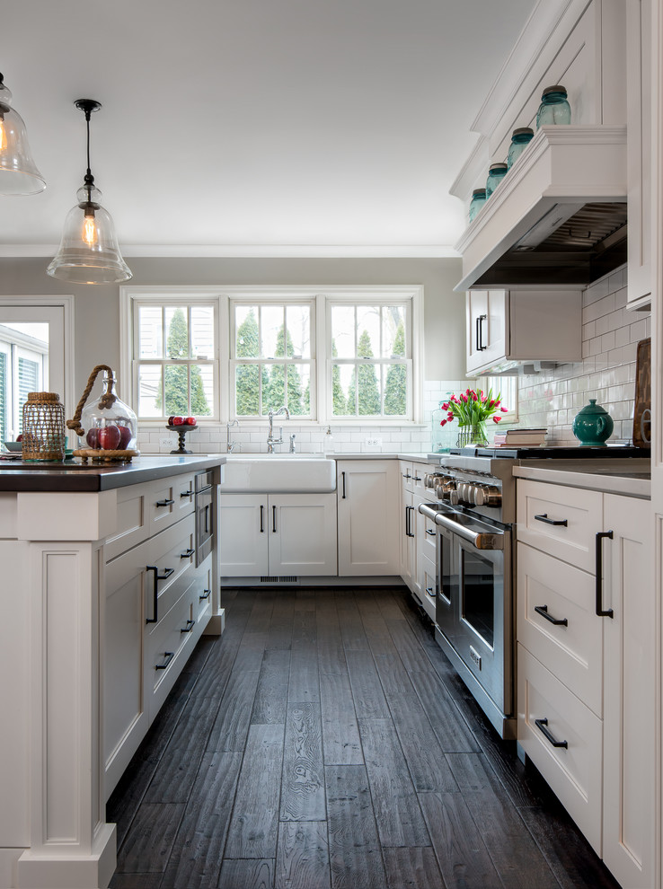 Photo of a large traditional l-shaped kitchen/diner in Detroit with shaker cabinets, white cabinets, white splashback, metro tiled splashback, stainless steel appliances, dark hardwood flooring, an island, a belfast sink, engineered stone countertops, brown floors and white worktops.