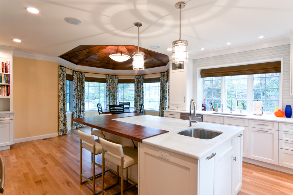 Inredning av ett klassiskt kök och matrum, med en undermonterad diskho, luckor med infälld panel, vita skåp, stänkskydd i glaskakel, rostfria vitvaror, träbänkskiva, vitt stänkskydd, ljust trägolv och en köksö