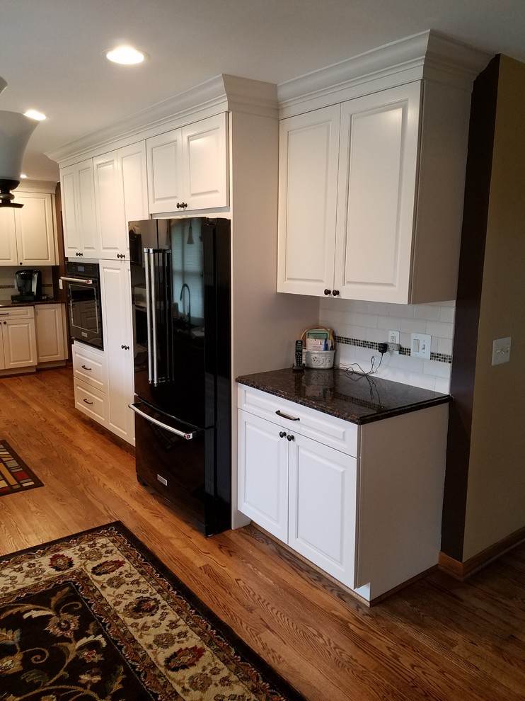 Eat-in kitchen - large traditional l-shaped medium tone wood floor and brown floor eat-in kitchen idea in Detroit with an undermount sink, raised-panel cabinets, white cabinets, granite countertops, white backsplash, ceramic backsplash, black appliances, an island and black countertops