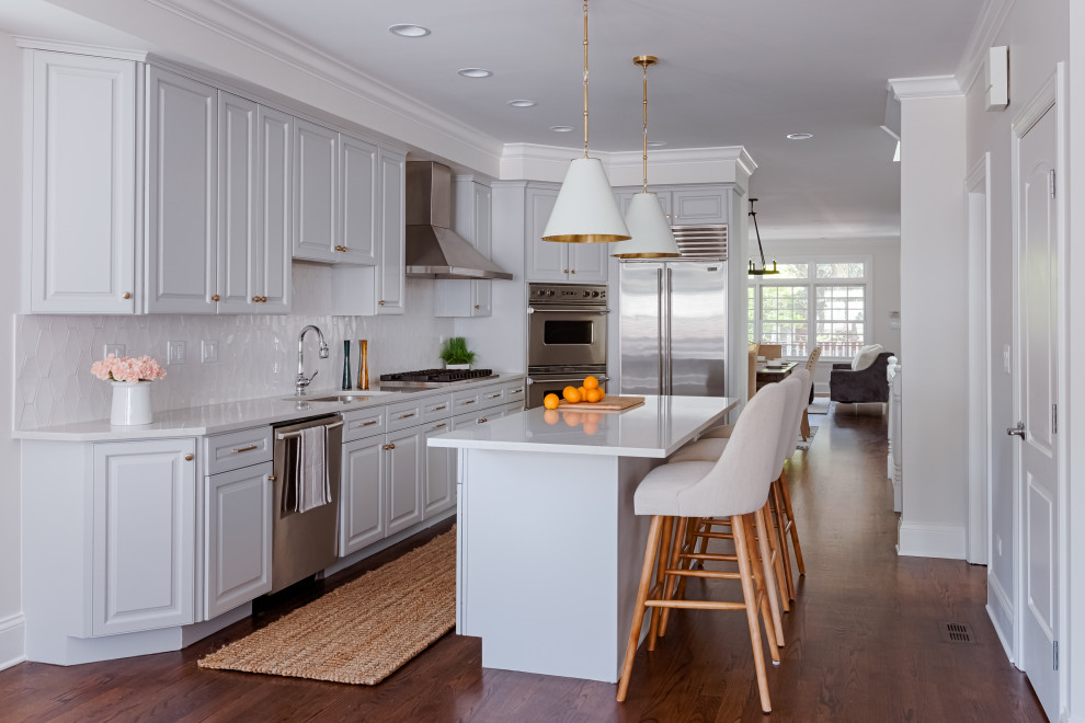 Klassisk inredning av ett vit vitt l-kök, med en undermonterad diskho, luckor med upphöjd panel, grå skåp, vitt stänkskydd, rostfria vitvaror, mörkt trägolv, en köksö och brunt golv