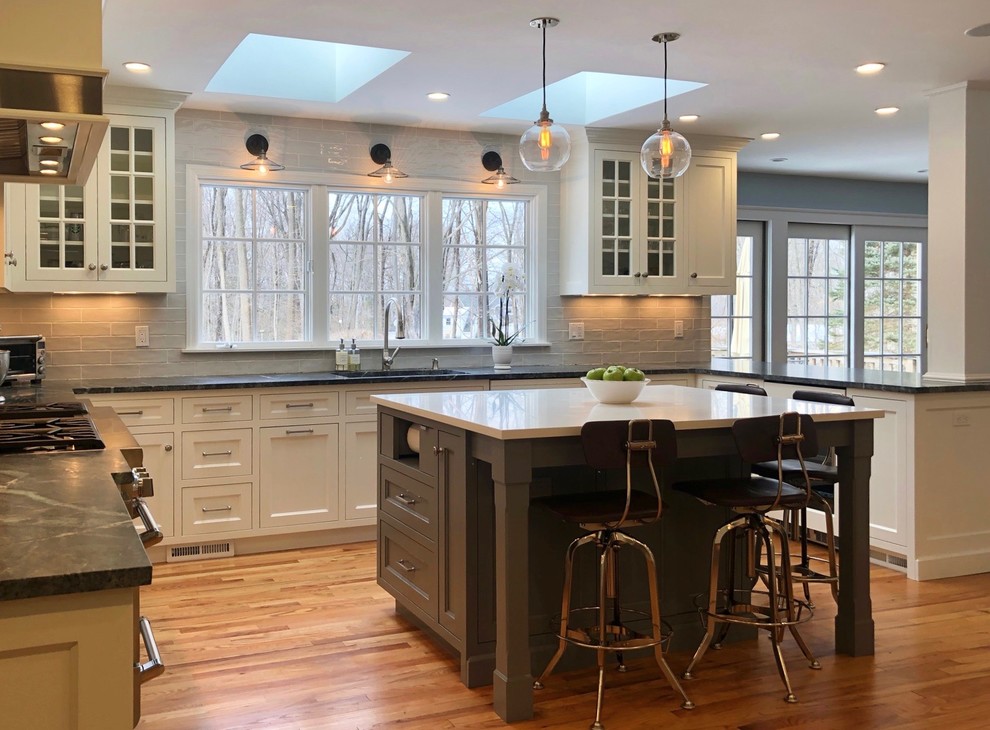 Inredning av ett klassiskt stort grå grått kök, med en undermonterad diskho, luckor med infälld panel, vita skåp, bänkskiva i täljsten, grått stänkskydd, stänkskydd i keramik, integrerade vitvaror, mellanmörkt trägolv, en köksö och brunt golv