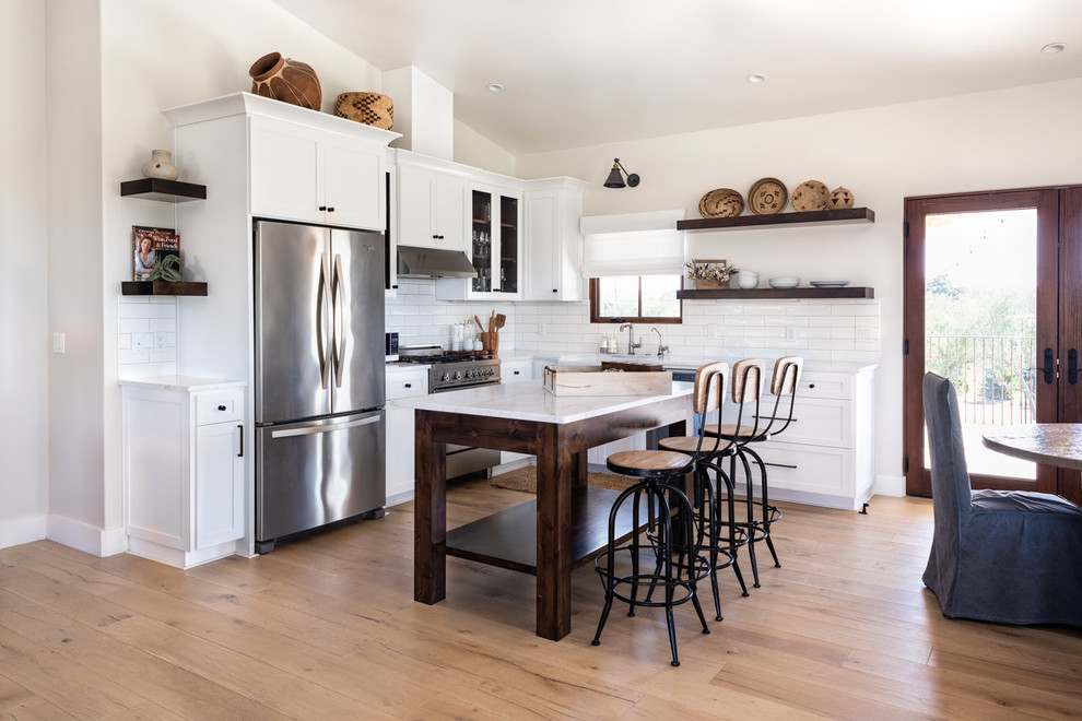 Inspiration for a mid-sized transitional l-shaped light wood floor and brown floor open concept kitchen remodel in San Luis Obispo with a farmhouse sink, shaker cabinets, white cabinets, marble countertops, white backsplash, subway tile backsplash, stainless steel appliances, an island and white countertops