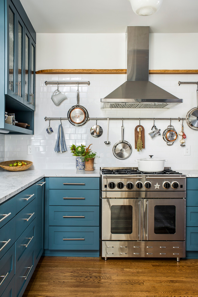 Bild på ett vintage l-kök, med skåp i shakerstil, blå skåp, vitt stänkskydd, stänkskydd i tunnelbanekakel, rostfria vitvaror och mellanmörkt trägolv