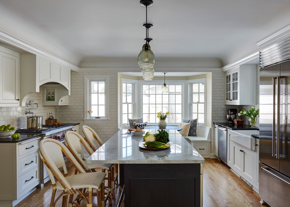 Klassisk inredning av ett kök, med en rustik diskho, luckor med infälld panel, vita skåp, bänkskiva i täljsten, vitt stänkskydd, stänkskydd i tunnelbanekakel, rostfria vitvaror, mellanmörkt trägolv och en köksö