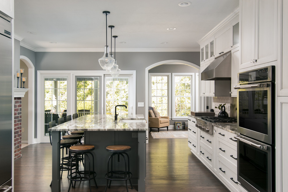 This is an example of a large classic grey and white kitchen in Cleveland with a belfast sink, recessed-panel cabinets, white cabinets, stainless steel appliances, dark hardwood flooring, an island, brown floors, metro tiled splashback, quartz worktops and white splashback.