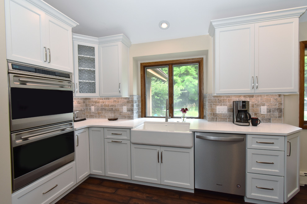 Example of a mid-sized transitional galley vinyl floor and brown floor enclosed kitchen design in Milwaukee with a farmhouse sink, recessed-panel cabinets, white cabinets, quartz countertops, beige backsplash, travertine backsplash, stainless steel appliances, an island and white countertops