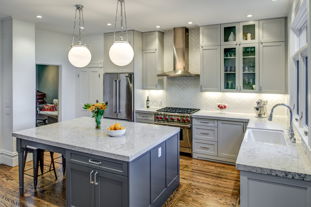 Enclosed kitchen - mid-sized transitional l-shaped medium tone wood floor enclosed kitchen idea in San Francisco with a farmhouse sink, raised-panel cabinets, gray cabinets, quartz countertops, white backsplash, porcelain backsplash, stainless steel appliances and an island