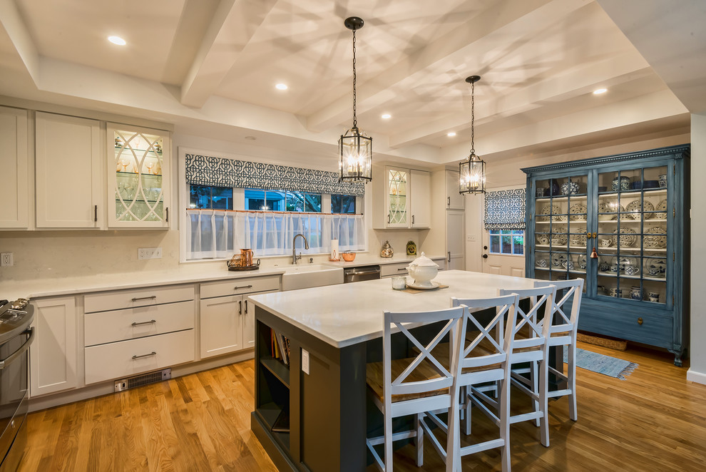 Example of a transitional l-shaped medium tone wood floor and brown floor kitchen design in Boston with a farmhouse sink, shaker cabinets, white cabinets, white backsplash, stainless steel appliances, an island and white countertops