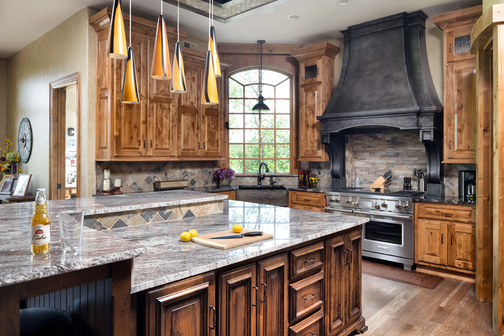 Open concept kitchen - large rustic l-shaped medium tone wood floor open concept kitchen idea in Kansas City with a farmhouse sink, raised-panel cabinets, brown cabinets, granite countertops, multicolored backsplash, stone tile backsplash, stainless steel appliances and an island