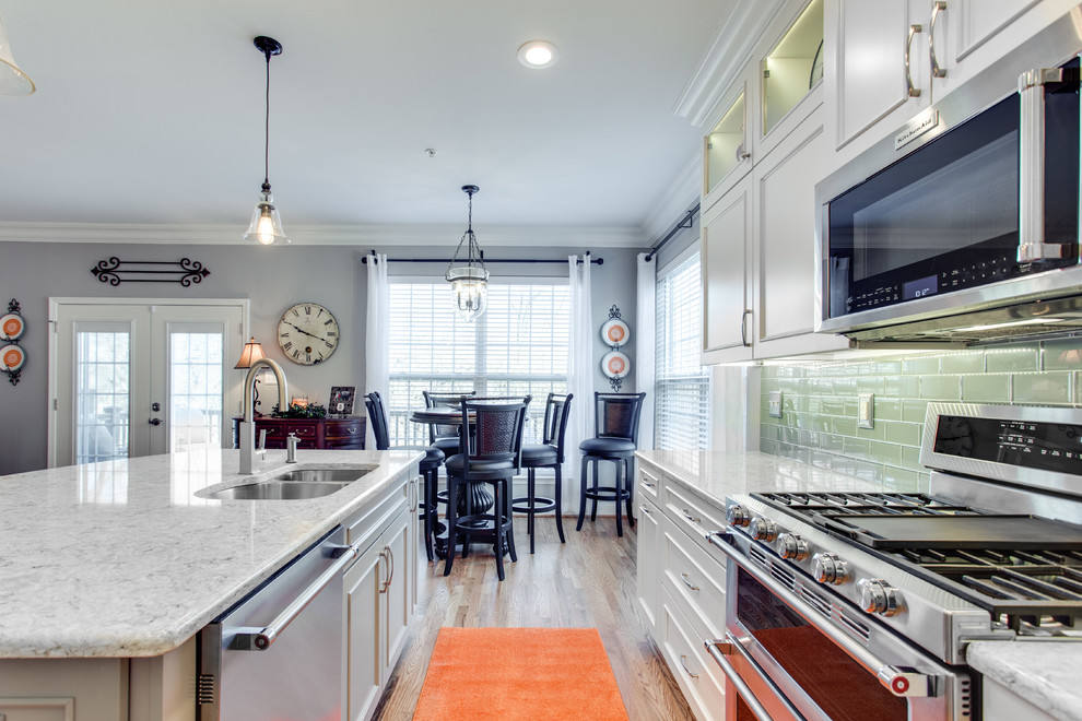 Mid-sized transitional l-shaped medium tone wood floor kitchen photo in Baltimore with an undermount sink, recessed-panel cabinets, white cabinets, quartz countertops, green backsplash, subway tile backsplash, stainless steel appliances and an island
