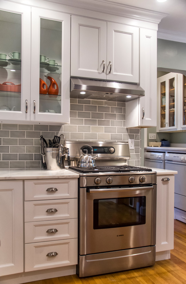 Example of a mid-sized transitional u-shaped medium tone wood floor and brown floor eat-in kitchen design in Bridgeport with a farmhouse sink, recessed-panel cabinets, white cabinets, marble countertops, gray backsplash, subway tile backsplash, stainless steel appliances and a peninsula