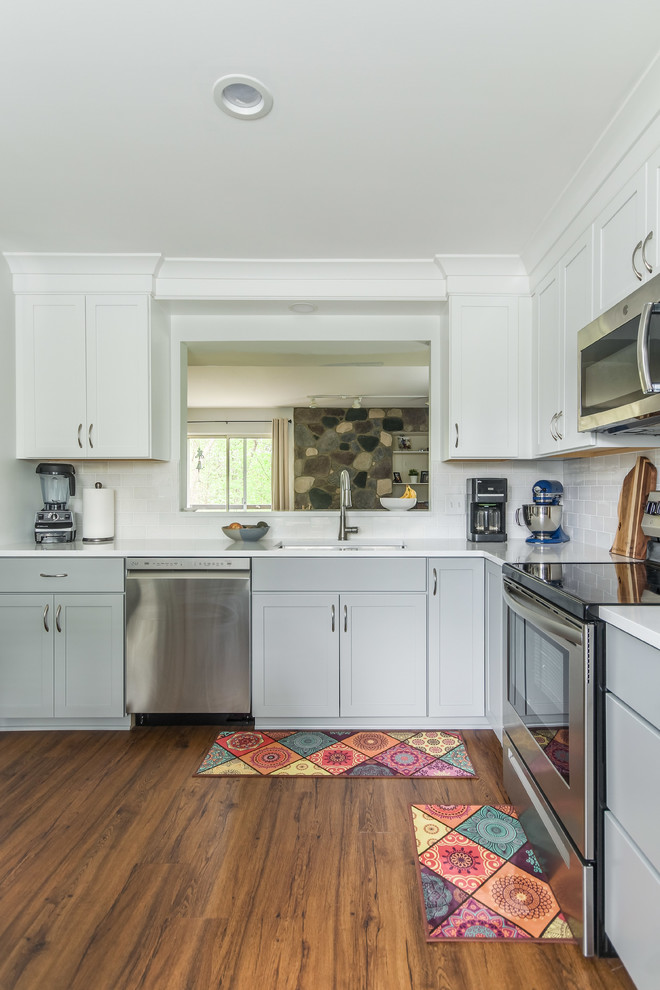Inredning av ett klassiskt litet vit vitt kök, med en undermonterad diskho, skåp i shakerstil, vita skåp, bänkskiva i kvarts, vitt stänkskydd, stänkskydd i tunnelbanekakel, rostfria vitvaror, mellanmörkt trägolv, en köksö och brunt golv