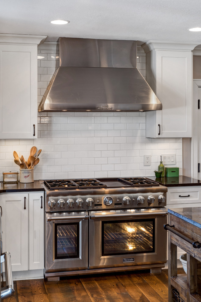 Foto på ett lantligt l-kök, med en rustik diskho, skåp i shakerstil, vita skåp, vitt stänkskydd, stänkskydd i tunnelbanekakel, rostfria vitvaror, mörkt trägolv och en köksö