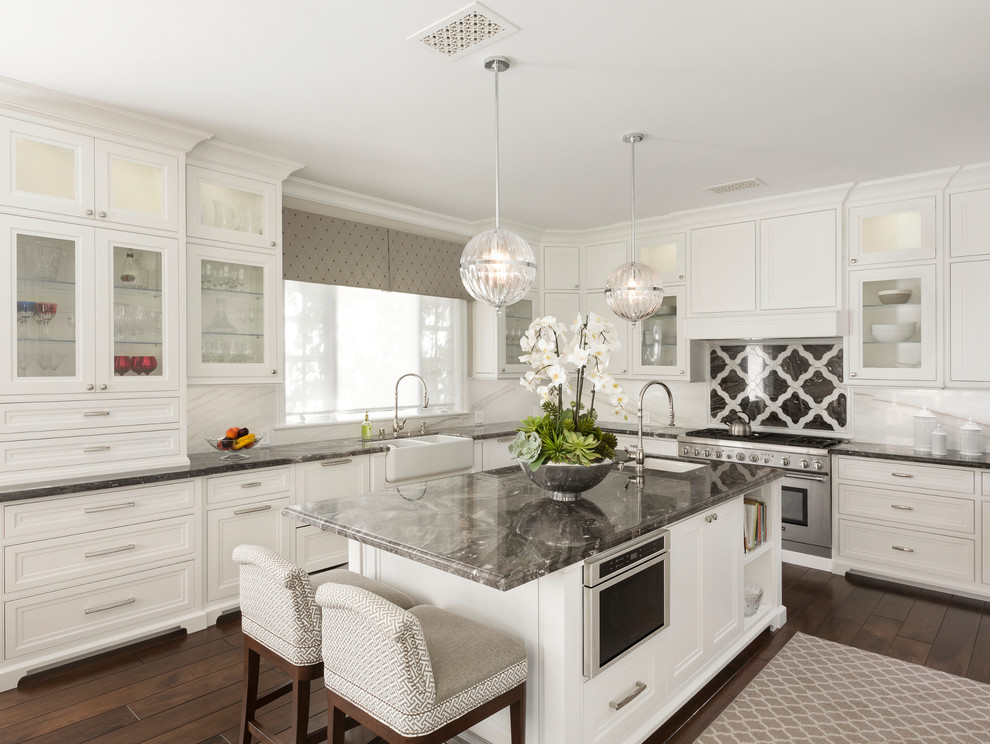 Example of a large classic l-shaped dark wood floor and brown floor enclosed kitchen design in Other with a farmhouse sink, glass-front cabinets, white cabinets, white backsplash, stainless steel appliances, an island, granite countertops, stone slab backsplash and gray countertops