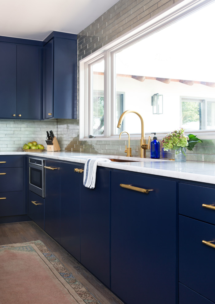 オースティンにある高級な中くらいなトランジショナルスタイルのおしゃれなアイランドキッチン (アンダーカウンターシンク、フラットパネル扉のキャビネット、青いキャビネット、大理石カウンター、メタリックのキッチンパネル、サブウェイタイルのキッチンパネル、シルバーの調理設備、淡色無垢フローリング、茶色い床、白いキッチンカウンター) の写真