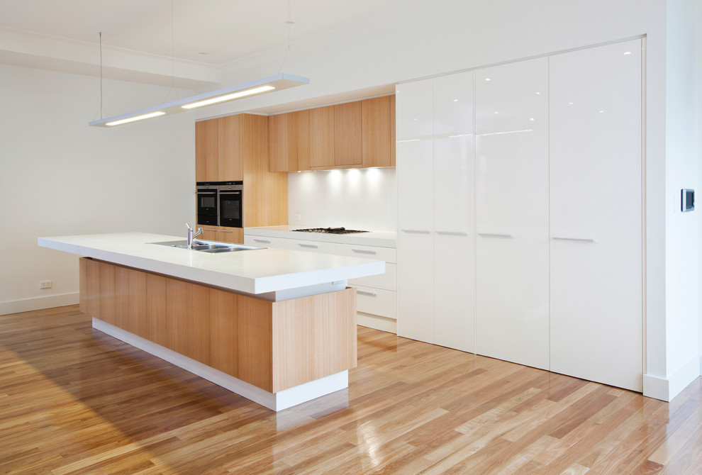 Inspiration for a large contemporary galley medium tone wood floor open concept kitchen remodel in Adelaide with a drop-in sink, flat-panel cabinets, light wood cabinets, quartz countertops, white backsplash, glass sheet backsplash, black appliances and an island