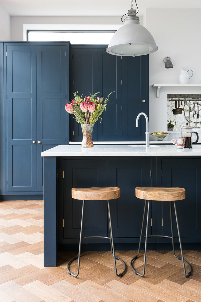 This is an example of a large traditional u-shaped kitchen/diner in London with a belfast sink, shaker cabinets, blue cabinets, marble worktops, metallic splashback, glass tiled splashback, integrated appliances, medium hardwood flooring and an island.